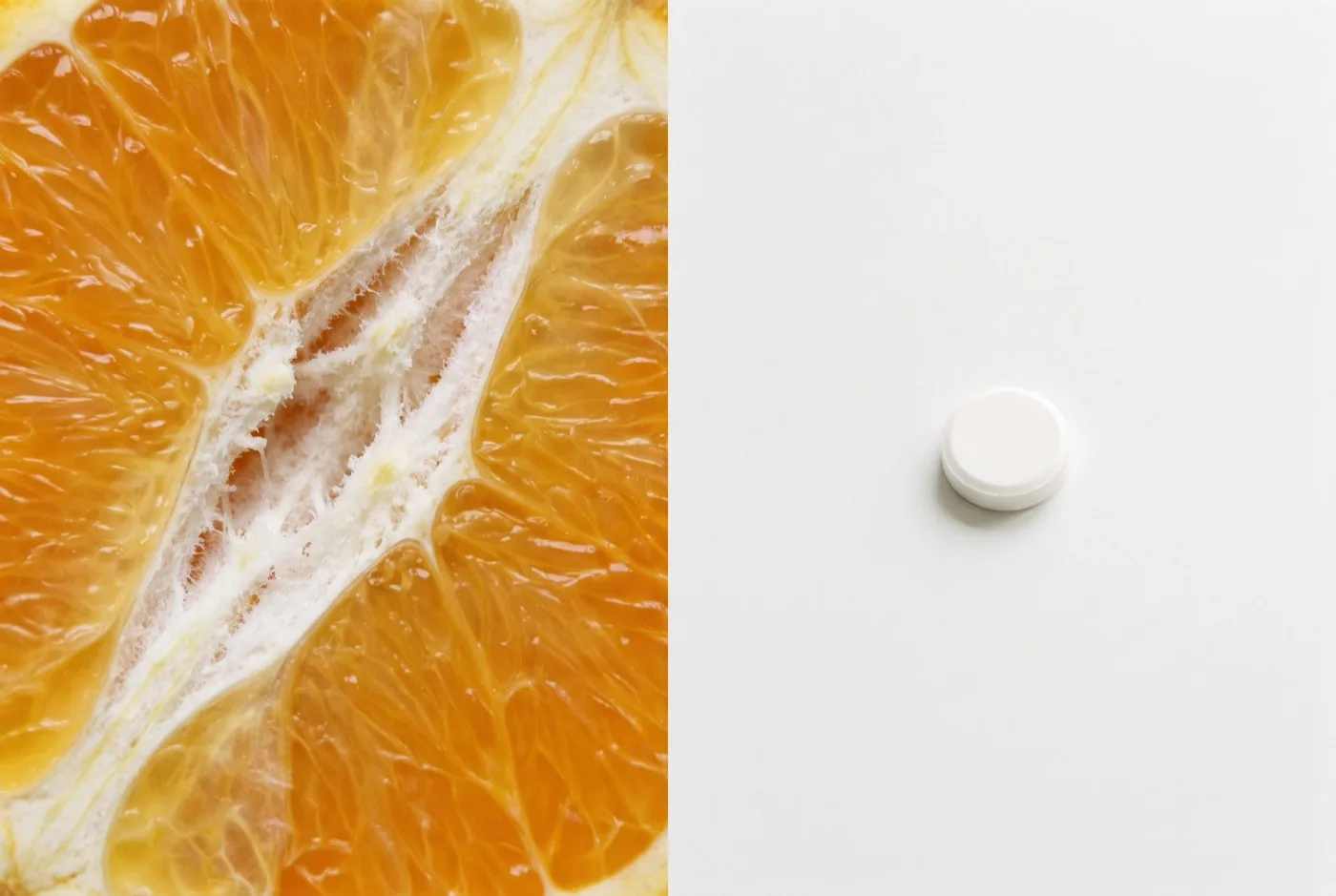 Extreme close-up of an orange segment with white pith beside a single isolated white vitamin C tablet on a plain surface