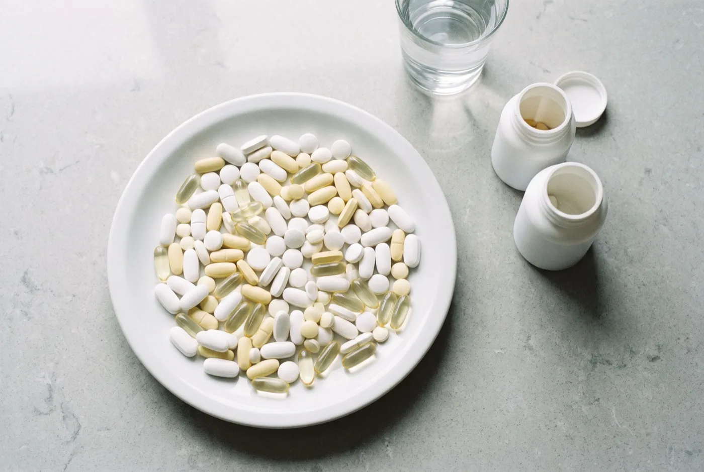 Overhead view of a white plate heaped with plain white and yellow tablets and softgels, two open unlabelled bottles and a glass of water