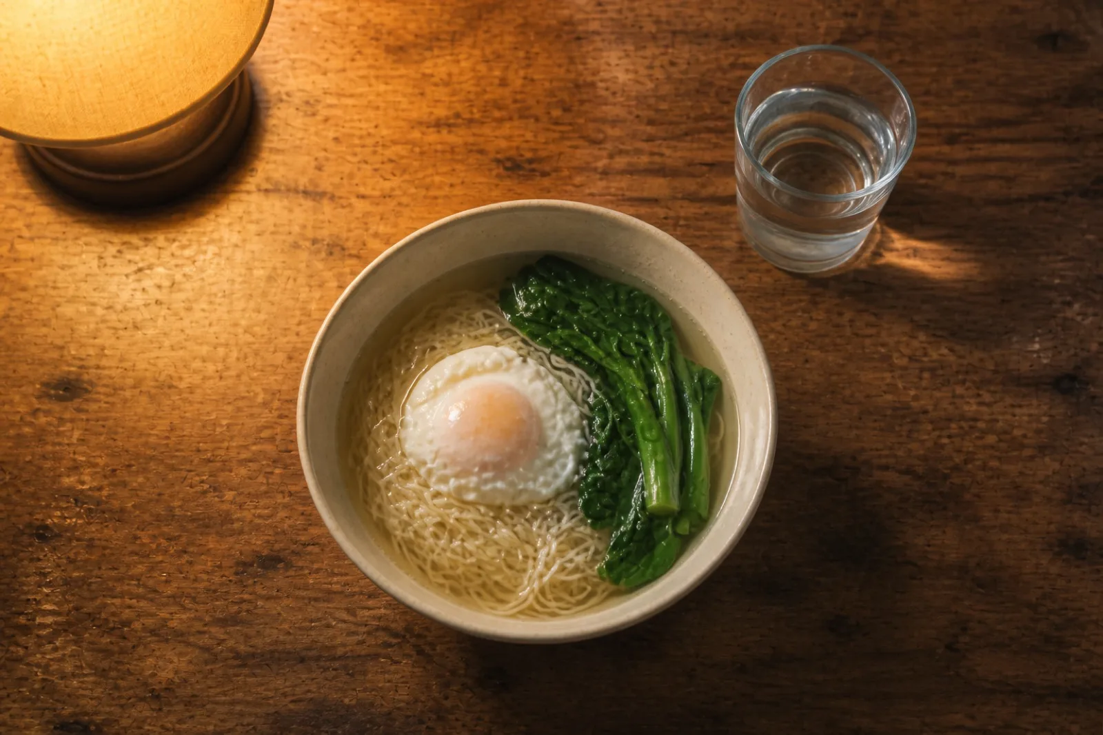 A simple late supper on a wooden table: a bowl of plain noodle soup with an egg and a few leaves of chye sim, a glass of water, warm lamp light